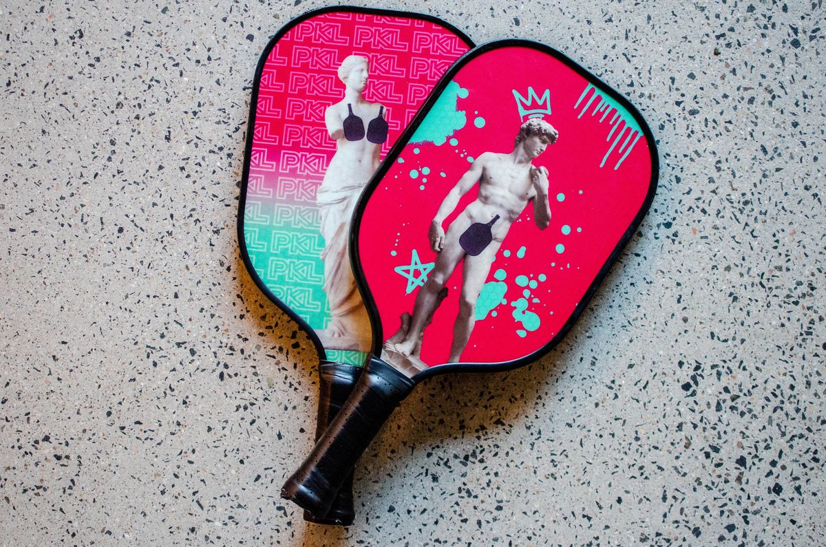 Two small pickleball paddles have a hot pink and light blue color scheme and feature historical nude statues with strategically placed pickleball paddles.