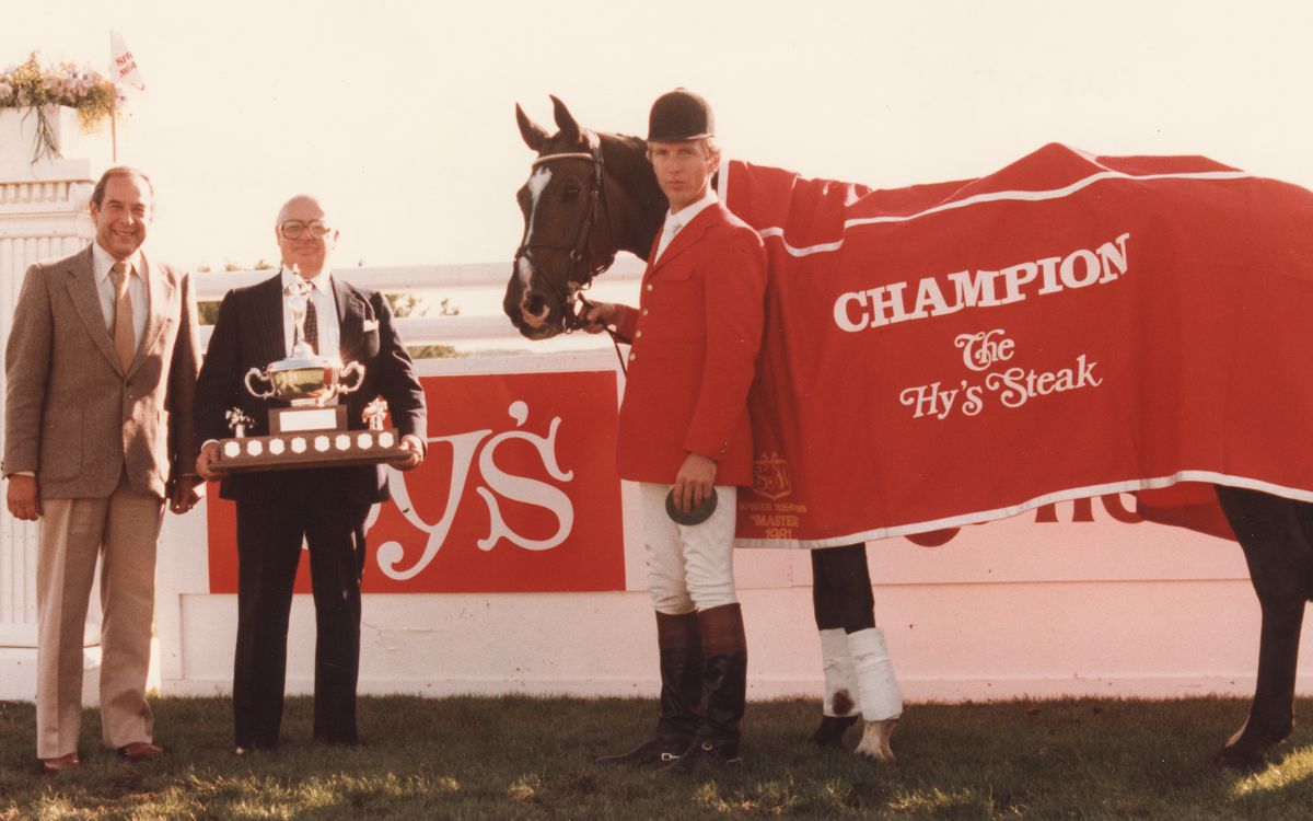 A jockey with a horse, caped in a red covering with the words Champion The Hy’s Steak, stand next to two men in suits, including Hy Aisenstat who holds a large trophy. 