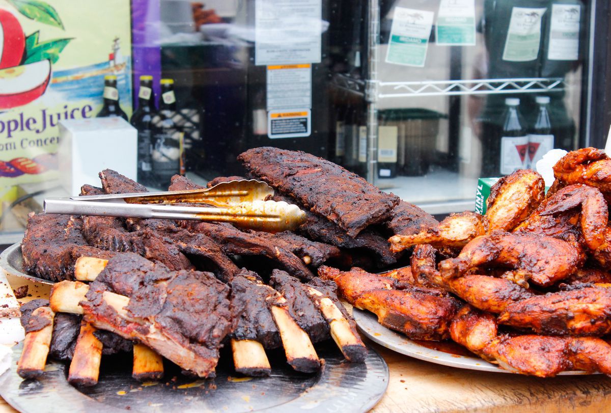 Platters of ribs and barbecue chicken sit on trays on a table outside of a shop window.