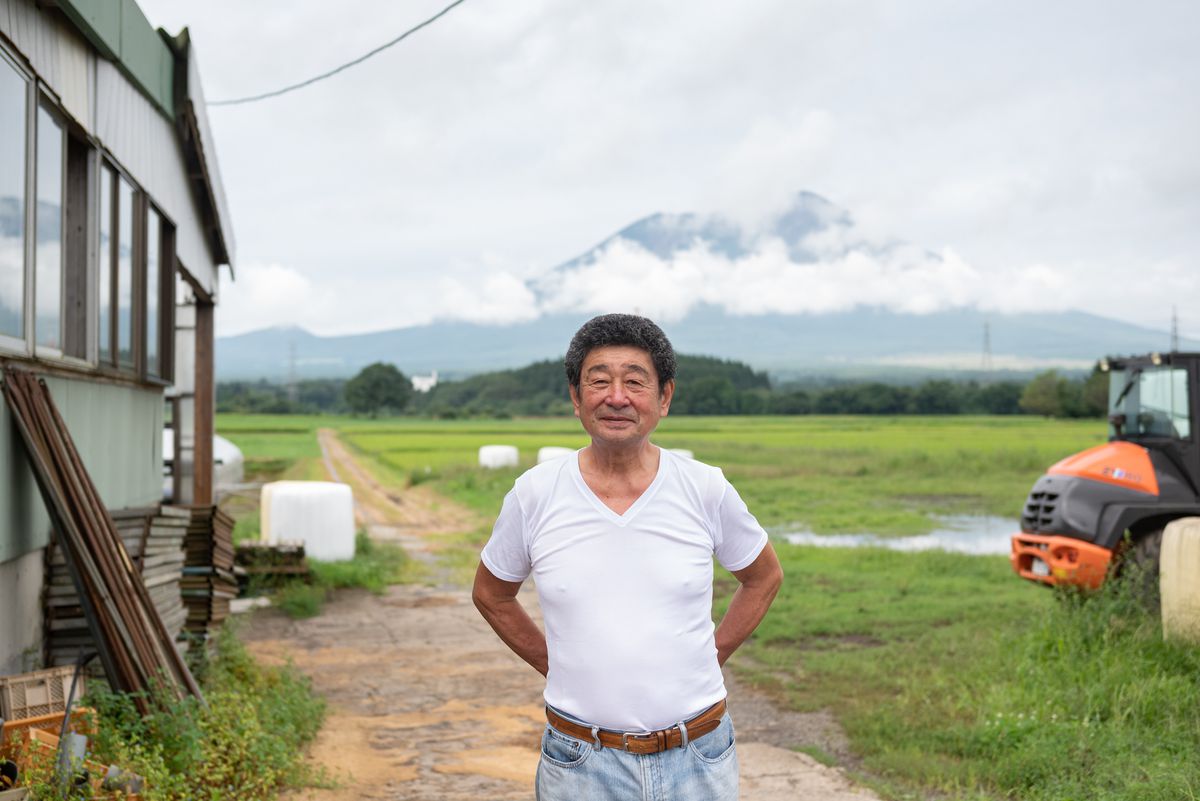 A farmer stands with his hands by his sides in front of a large green expanse, with cloud-covered mountains beyond.