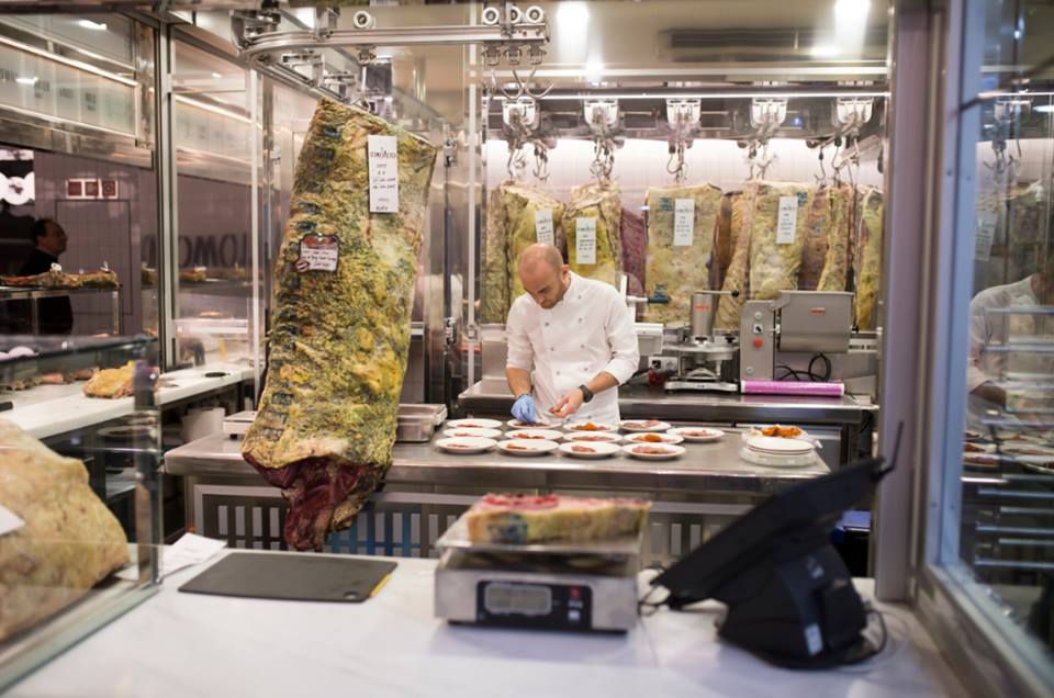 A chef working in a butchery room surrounded by hanging meat