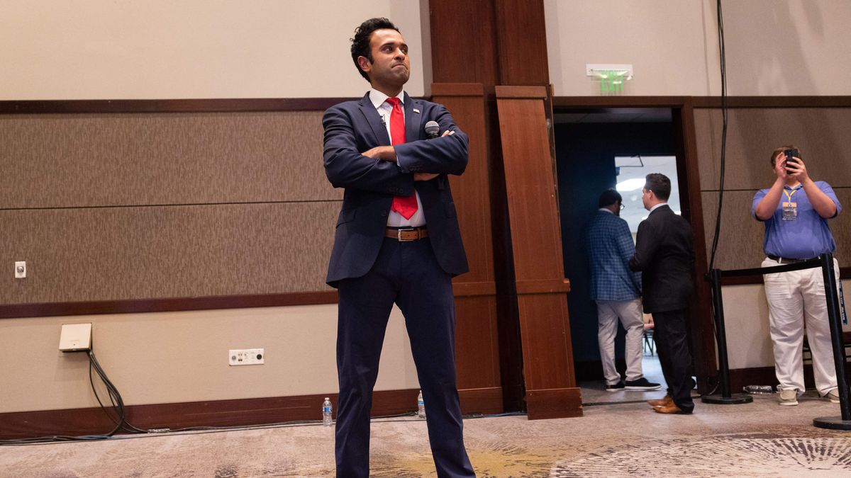 Vivek Ramaswamy stands in a ballroom, wearing a suit, arms crossed.