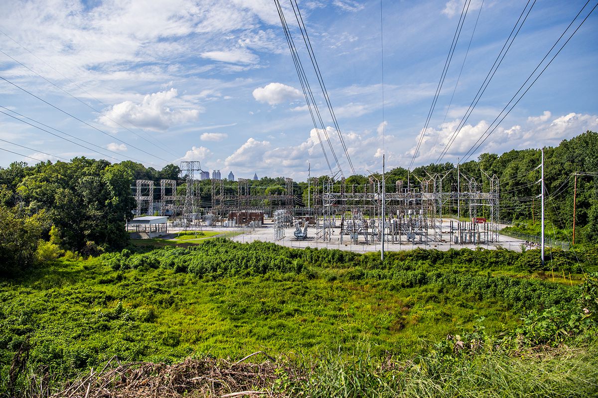 A power substation is shown with power lines overhead and a skyline in the distance. 
