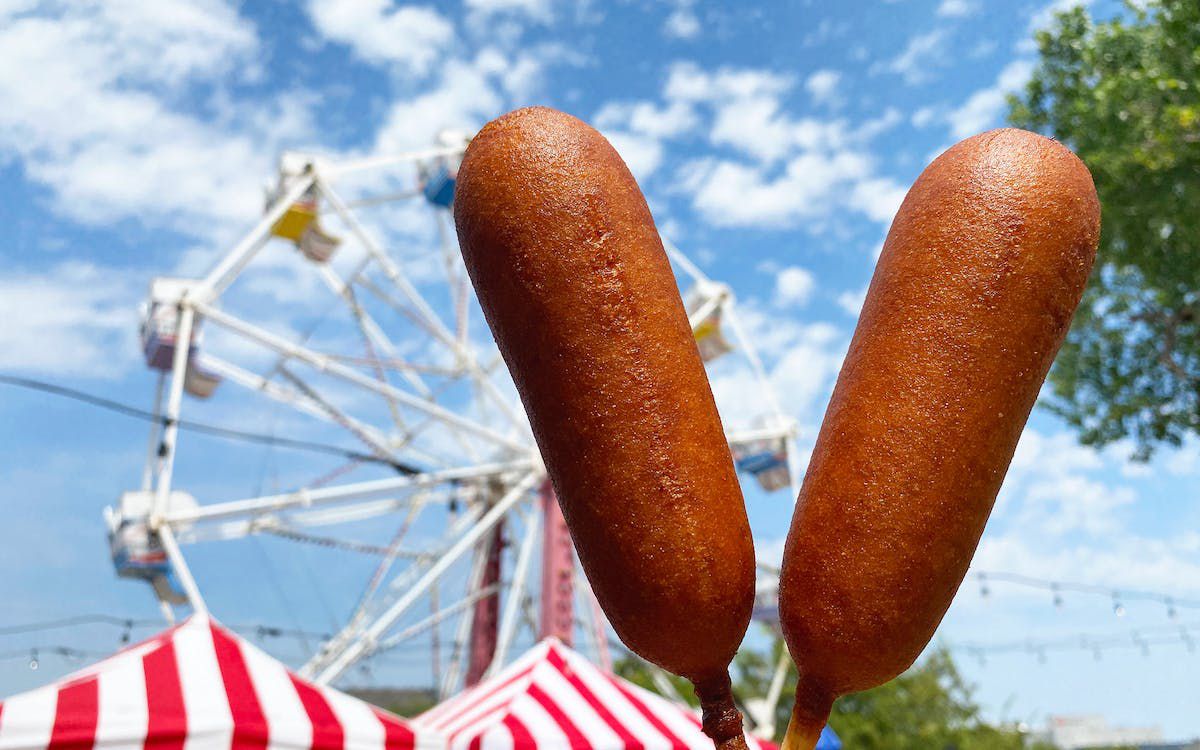 Two corn dogs are held by one hand, with a circus tent and blue sky in the background. 