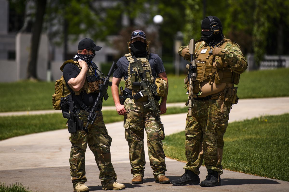 Three people in camouflage clothing stand with rifles and other gear in their hands and slung on their shoulders.