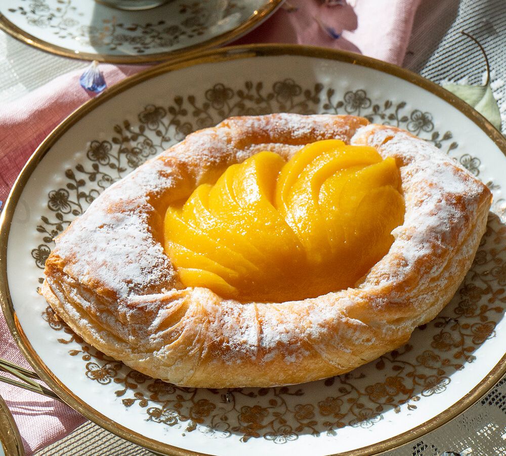 A diamond-shaped danish filled with bright yellow slices of peach, the edges dusted with sugar, on a decorative plate beside an ornate mug of tea