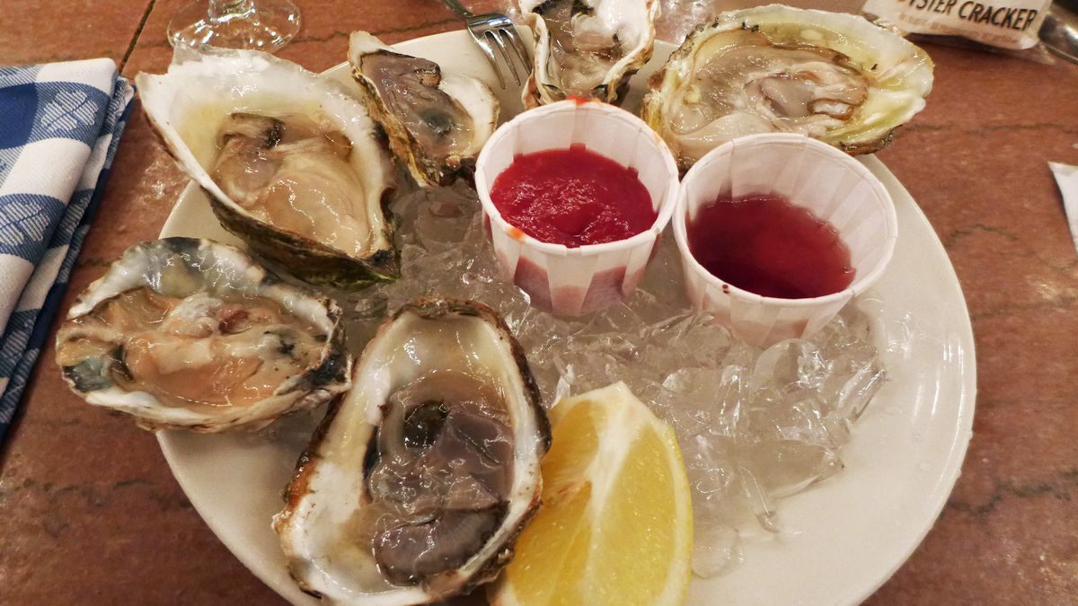 Six oysters of different shapes and sizes, on a plate lined with ice, served with a lemon wedge...