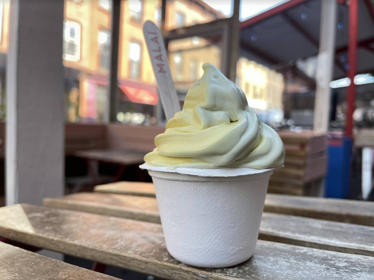 A white paper cup with a wood spoon is filled with yellow and white swirled soft serve.