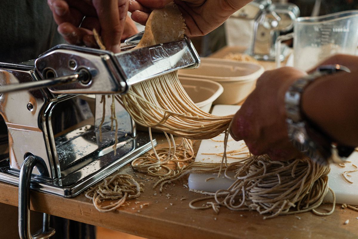 Ramen noodles are cranked out of a pasta machine into a pair of waiting hands.