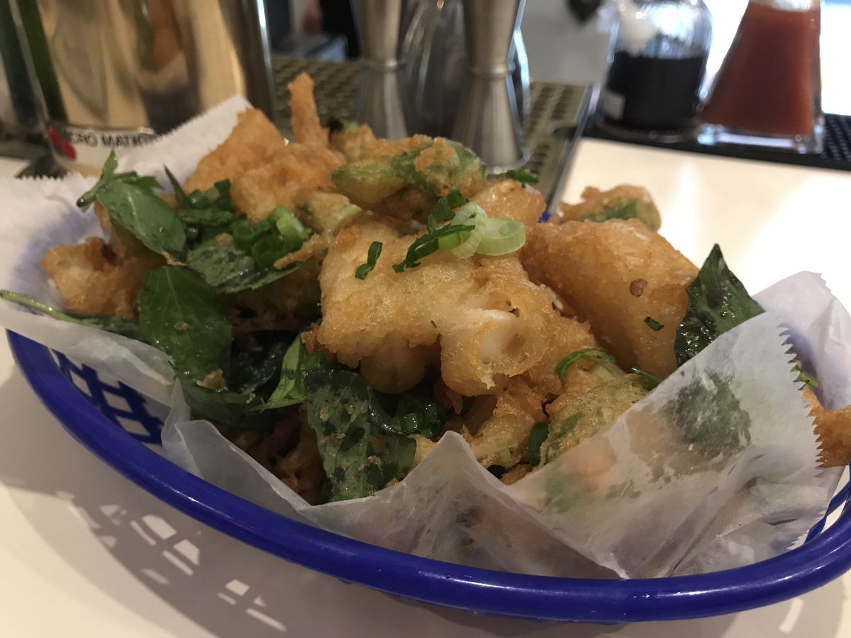 A blue plastic basket with tissue paper, filled with fried calamari, fried basil, and fried shishito peppers