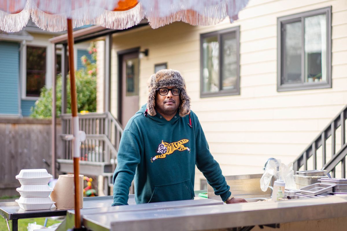 A man stands behind a flat-top of a food cart wearing a teal sweater with a yellow tiger on it and a fur trapper hat.