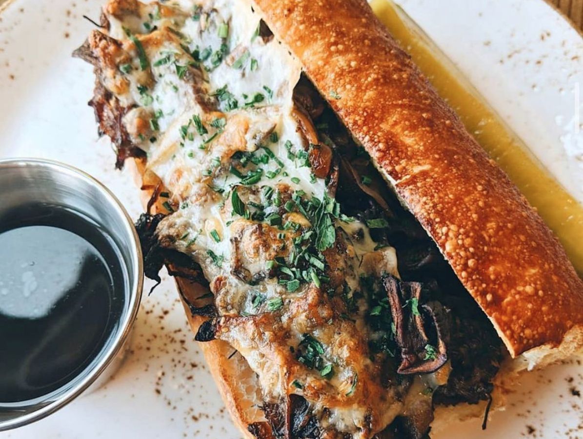 The oxtail french dip sandwich shows piled oxtail with melted while havarti cheese, charred onions, and mushrooms on a toasted bread roll. On the plate, in a small silver ramekin, is rosemary jus sauce for dipping.