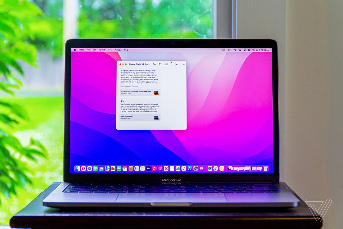 A MacBook Pro running macOS Monterey sits on a table in front of a window. The screen displays the Quick note app on top of a pink and purple background.
