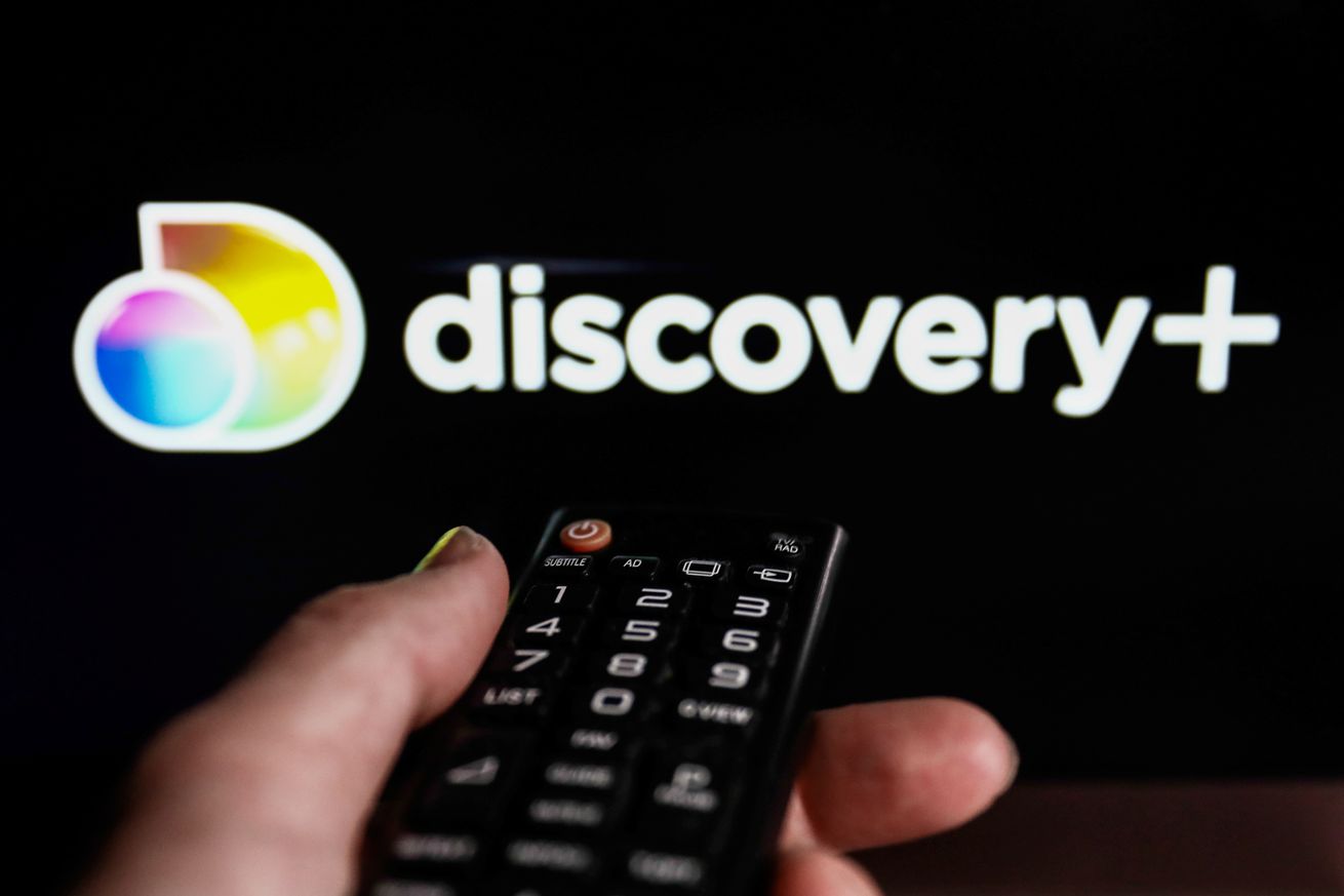 An image showing a remote control pointed on a screen that has the Discovery Plus logo