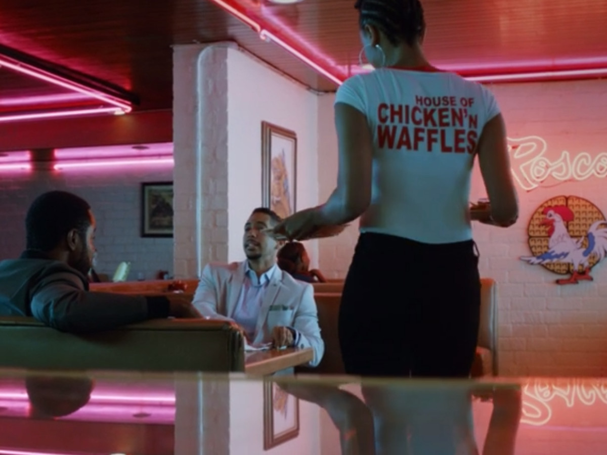 The interior of a restaurant. There are neon signs and decorative neon elements. There are two men sitting at a table talking. A waitress is bringing the men food. Her shirt reads Roscoe’s House of Chicken and Waffles. 