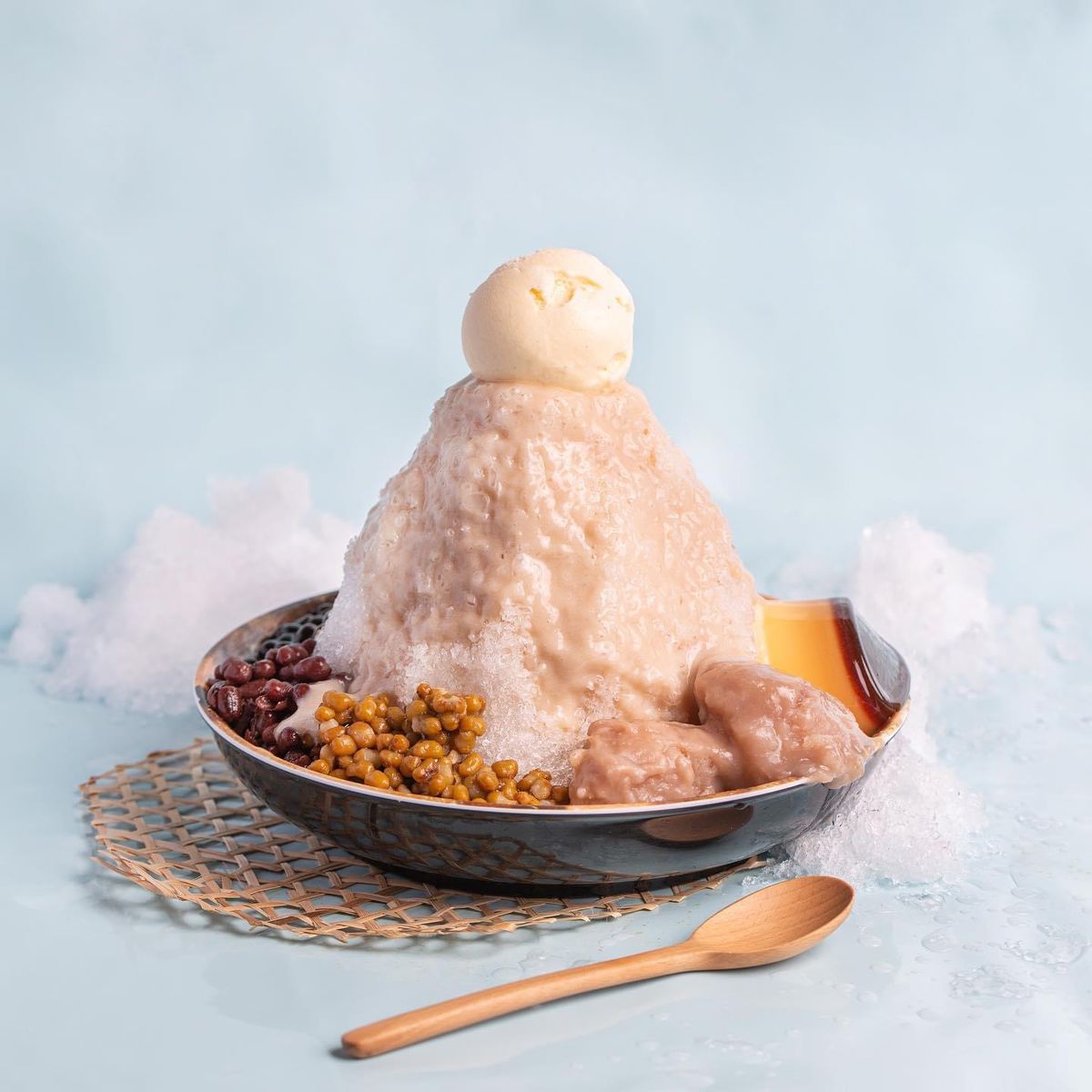 A tall serving of shaved ice is served with sweetened beans, brown sugar boba, and topped with a scoop of pudding.