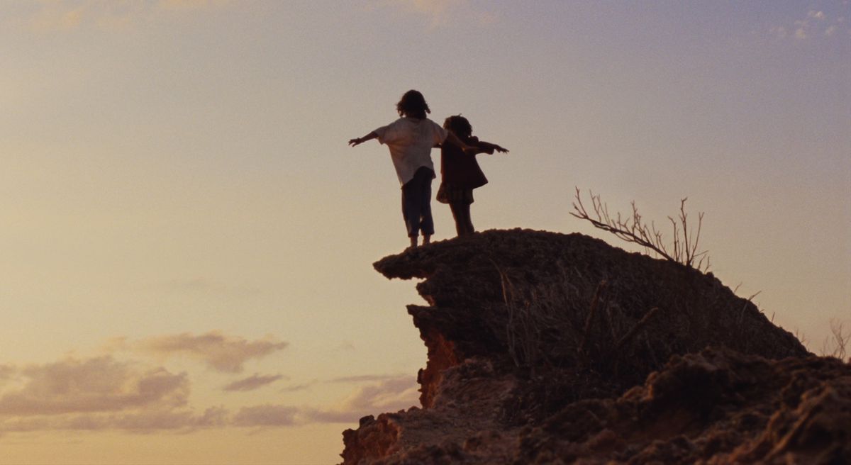 A girl and a boy stand on a barren reddish rock cliff at sunset, arms out as if they intend to fly, silhouetted against a mostly empty sky.