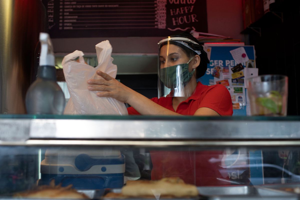 Woman wearing face mask and face shield preparing plastic bag of food from behind a counter.