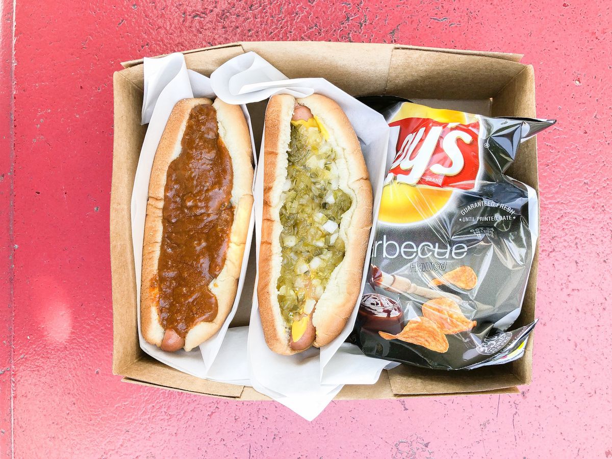 Two hot dogs with sauce and relish next to chips on a red table.