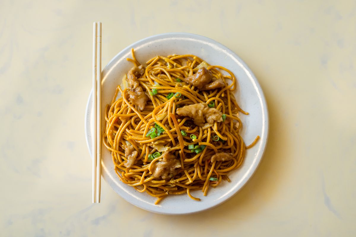 A white plate of chicken lo mein sits on a beige tablecloth.