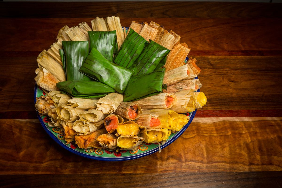 A platter of tamales stuffed with meat, with seven tamales wrapped on top with a green banana leaf.