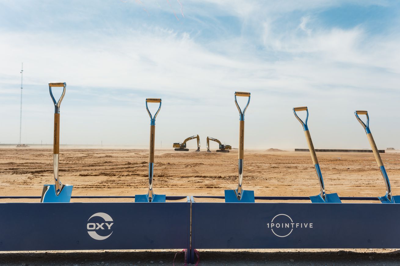 A row of five shovels standing up behind signs with logos for Occidental Petroleum and 1PointFive. Construction equipment can be seen in the background.