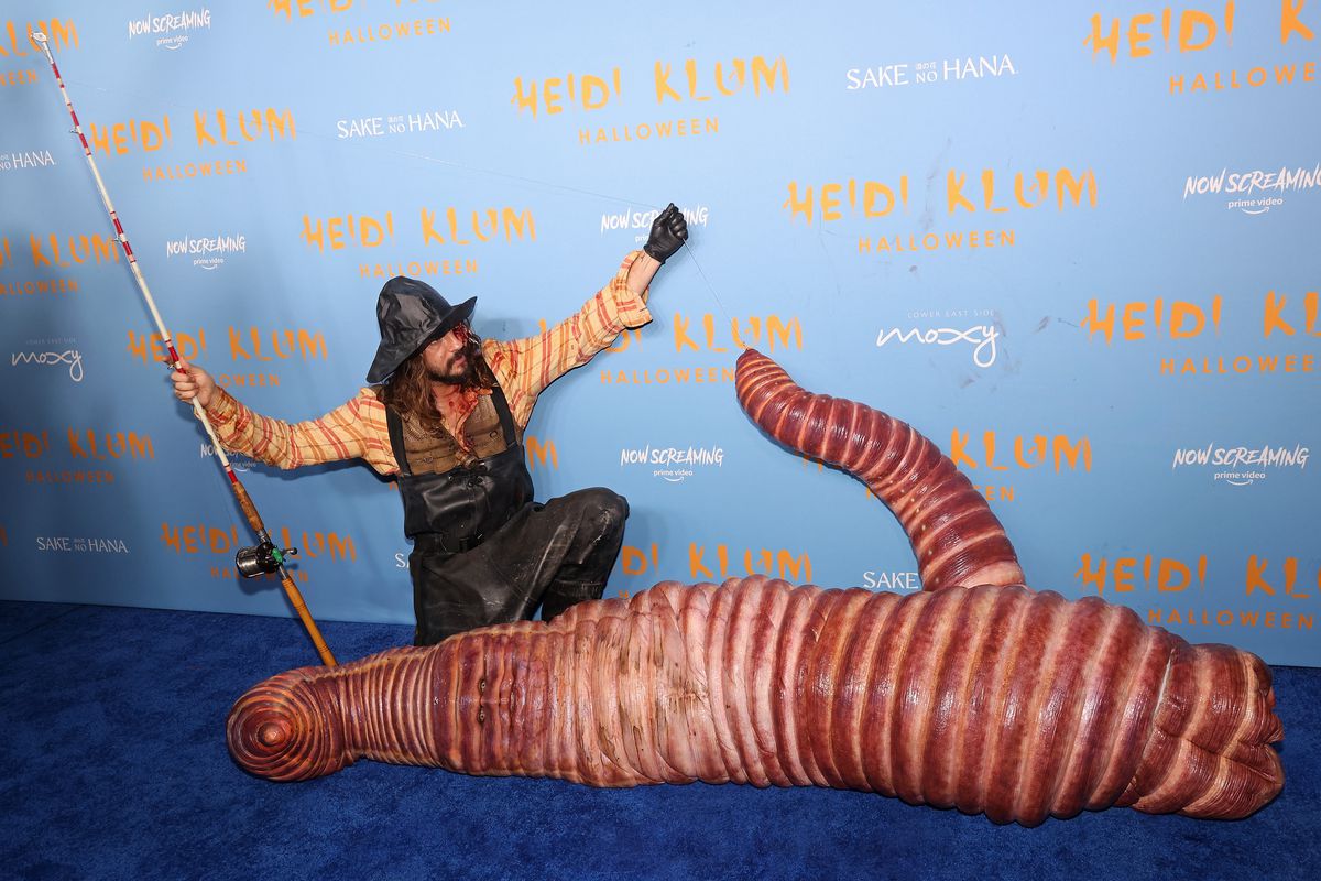 A photograph of the German American model Heidi Klum and her husband Tom Kaulitz dressed in their Halloween costumes at Klum’s Hallowe’en Party 2022. Klum wears an eerily realistic worm costume and is laying down on the ground — No but seriously, it looks super fleshy and gross. 