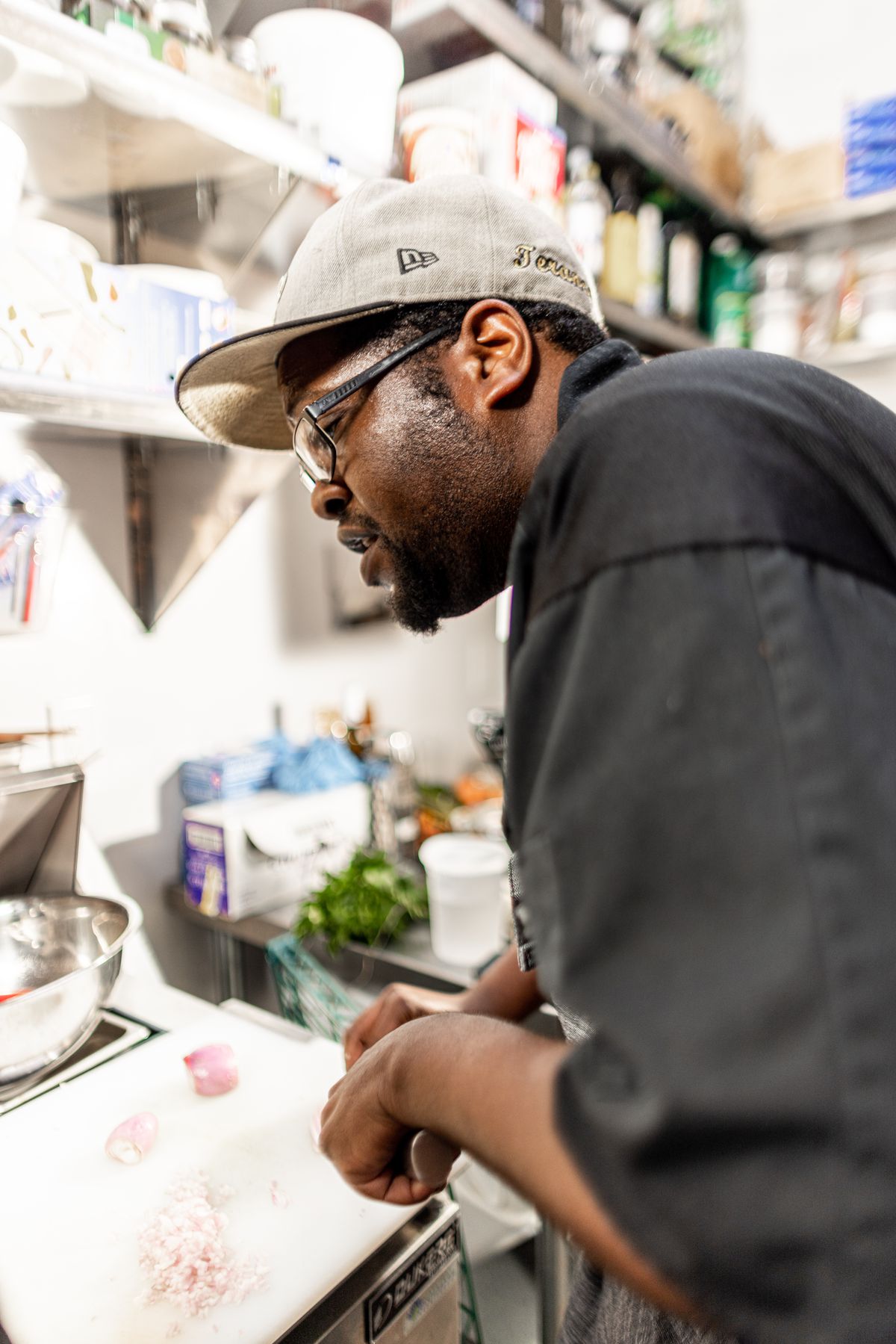 Chef Terance Jenkins, a man in a baseball cap, black-rimmed glasses, an apron, and black chef’s clothes, preparing food in the kitchen at Restaurant Beatrice.