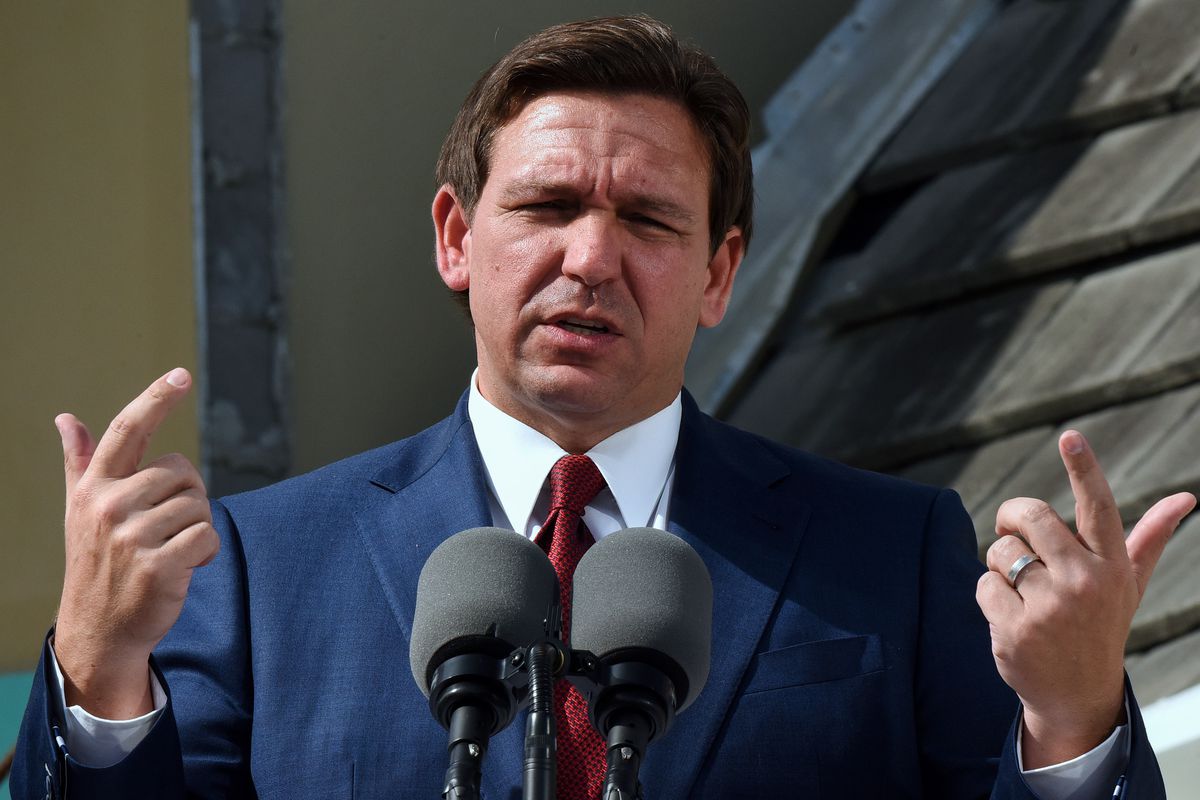 Ron DeSantis speaks in front of microphones, making a gesture with both hands, pointer fingers and thumbs extended.