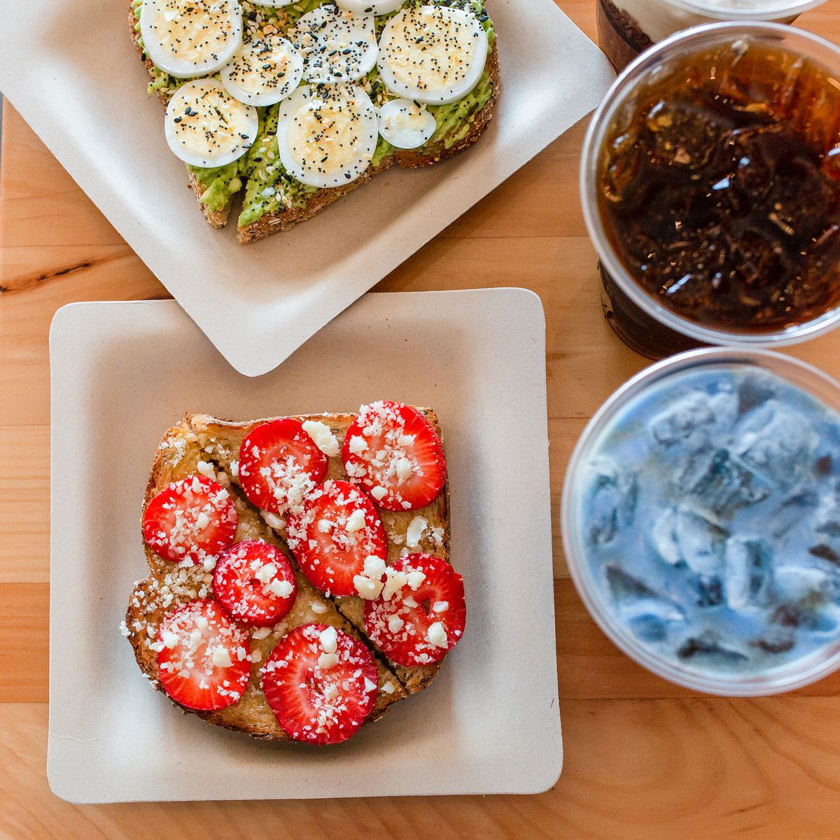 Plated avocado toast and strawberry toast and two beverages with ice, one brown and one blue