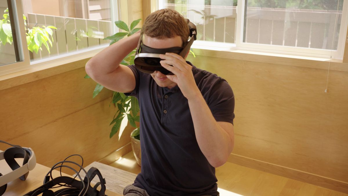 Mark Zuckerberg trying on a black VR headset prototype, surrounded by other headsets