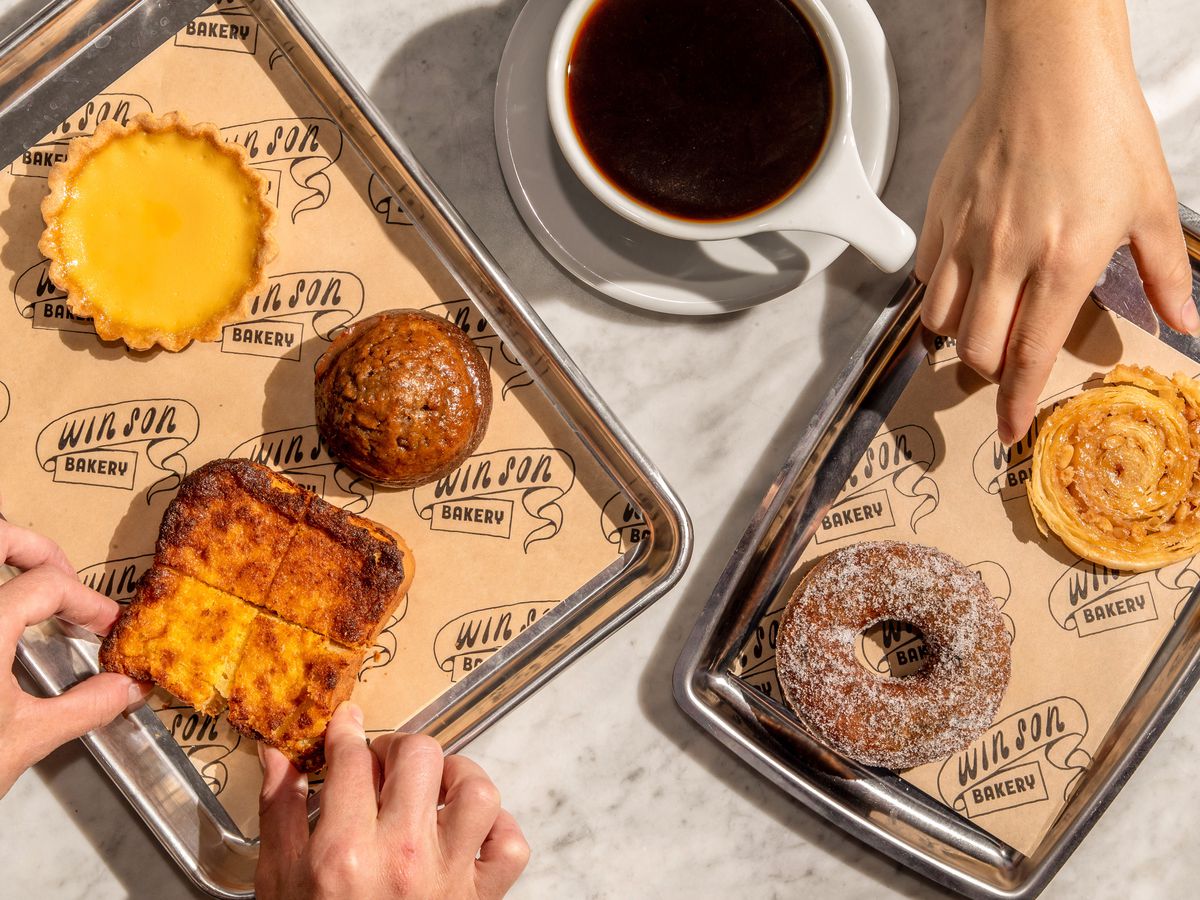 Assorted pastries, including mochi doughnuts and bright yellow custard toast, on two stainless steel trays alongside a cup of coffee, at Win Son Bakery