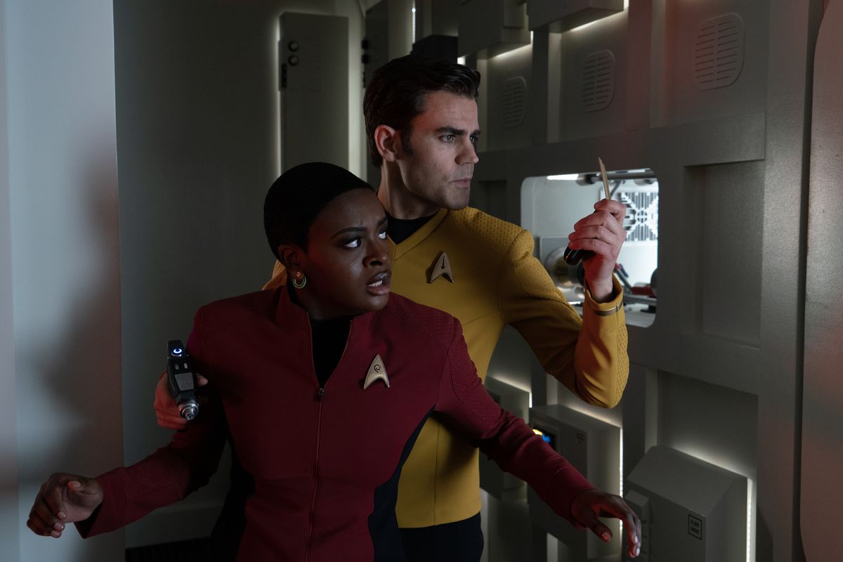 Uhura (Celia Rose Gooding) standing in front of Kirk (Paul Wesley) who’s checking his transponder