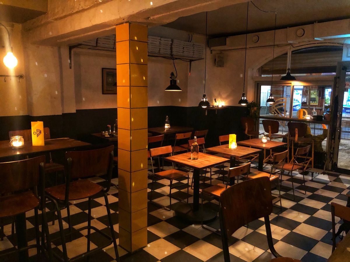 A restaurant interior lit by soft pendants, with checkered floor tiles, low and high tables, exposed cement ceilings, and a single tiled column rising through the center of the room