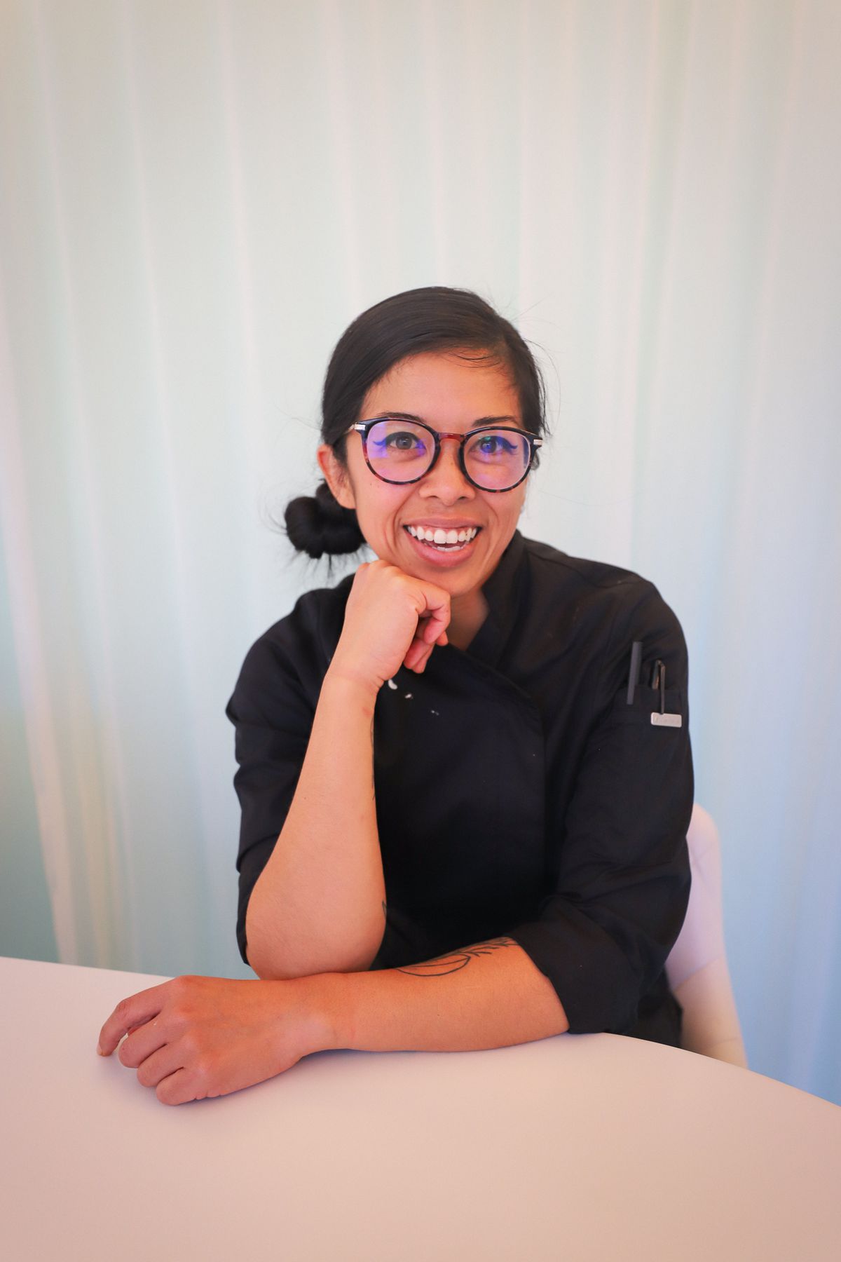 A woman in a black chef’s coat with glasses and hair tied back in a low bun sits at a table and smiles at the camera.