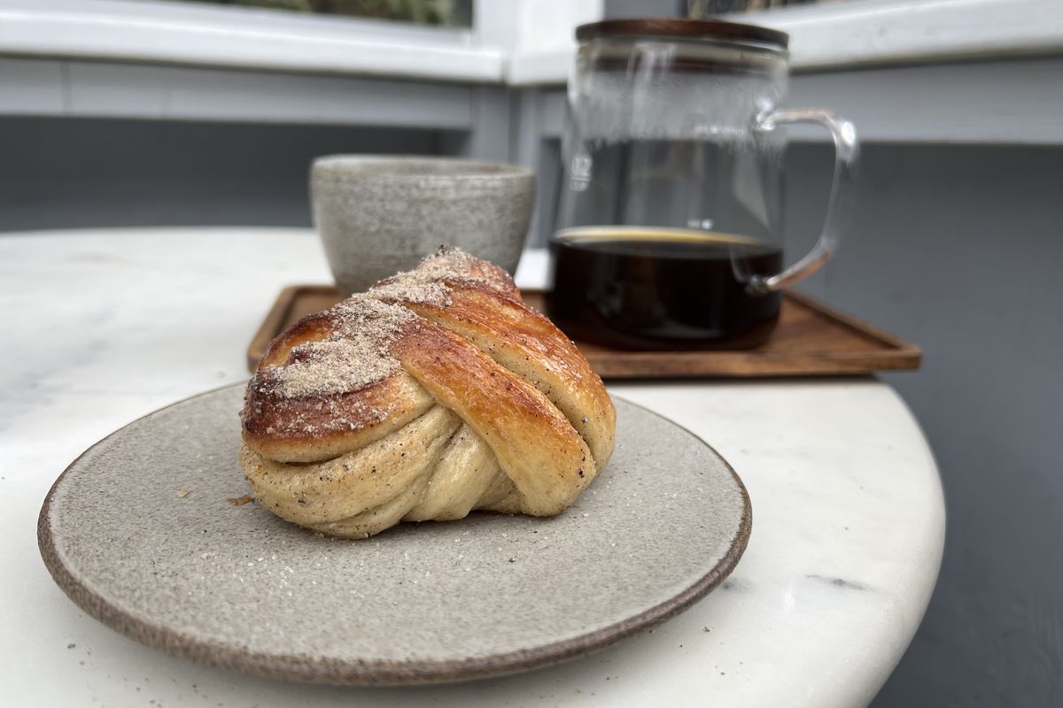 A cardamom bun sits on a grey plate on a white table, just in front of a clear coffee flask and an earthenware cup