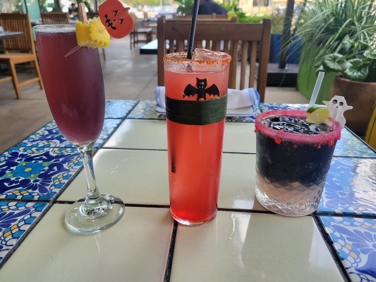A cocktail in a champagne glass with a pumpkin garnish, a cocktail in a high ball glass with a bat on it, and black-colored cocktail with a pink rim and ghost-shaped garnish.