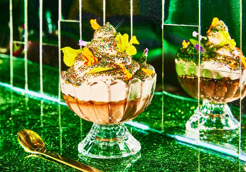 A tall swirl of ice cream in a sundae dish topped with sprinkles and other bright accoutrements, sitting on a glittery green bar in front a mirrored wall.