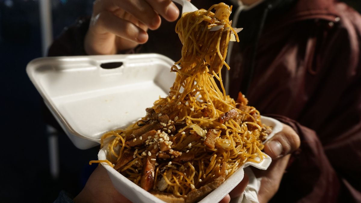 A hand holding a pair of chopsticks suspends a heap of noodles above a Styrofoam takeout tray