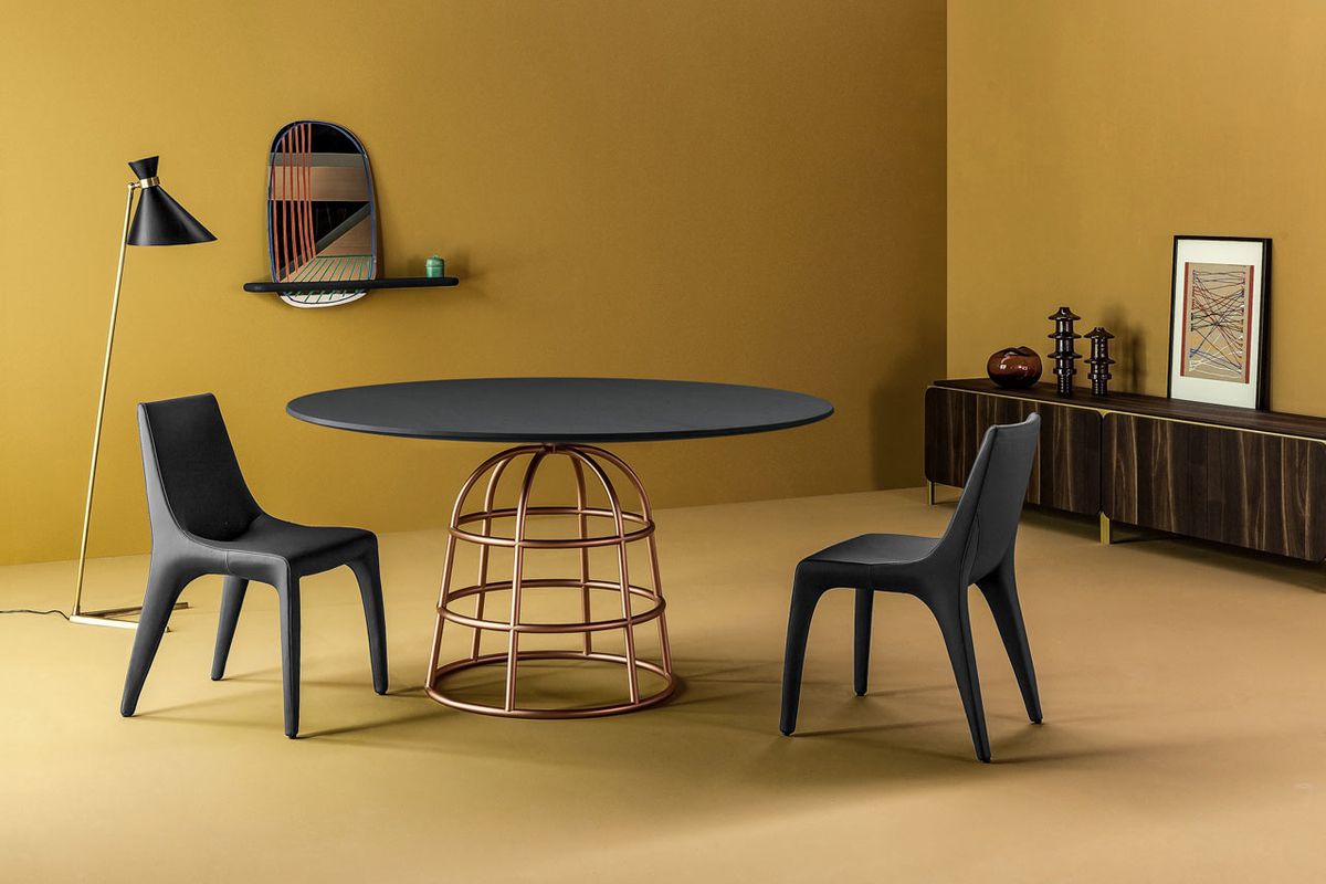 Showroom shot of a dark round table with a gold, metallic base in the shape of a birdcage surrounded by two black plastic chairs in a mustard-colored room. A conical floor lamp and a low credenza complete the scene.