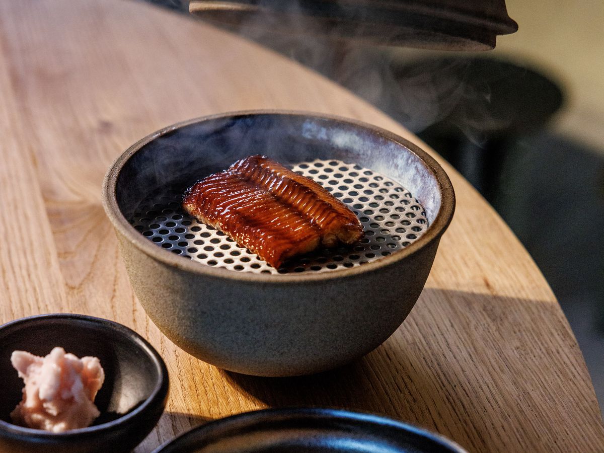 A lid lifts off a mini grill revealing glazed eel, presented beside pickled vegetables and another blurred dish. 