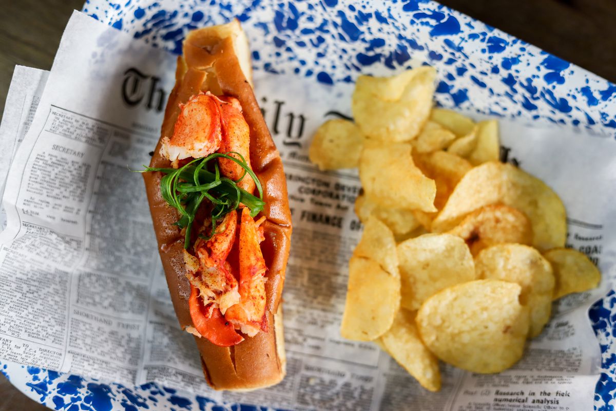 A lobster roll and potato chips on newspaper on a blue speckled plate.