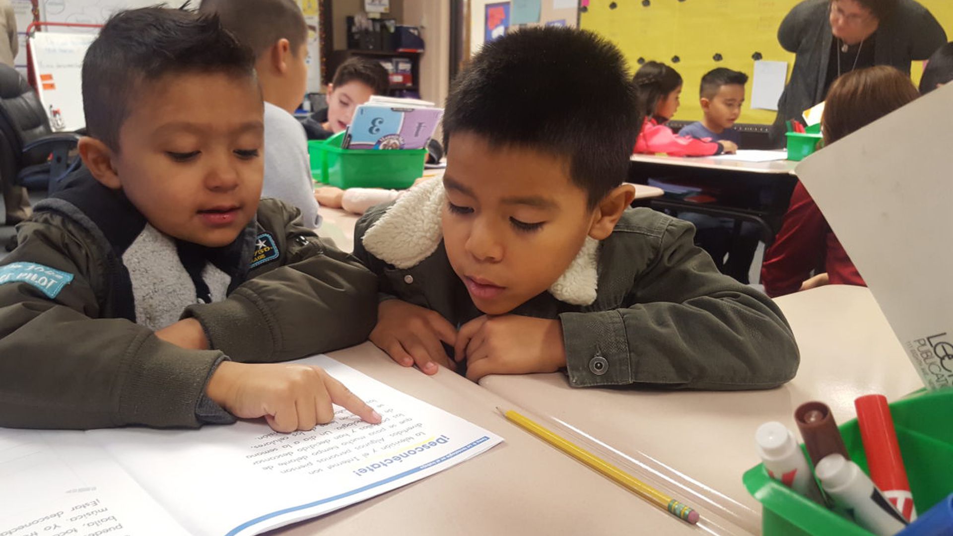 Two first grade boys look at a reading workbook at a desk in a classroom.