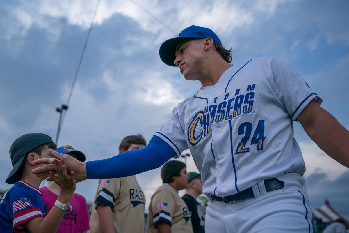 Picture from a low angle of a man in a white baseball uniform and blue cap giving high fives to a line of children.