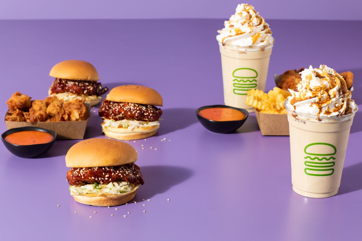 An arrangement of fried chicken sandwiches, chicken nuggets, fries, sauces, and two milkshakes on a purple background.