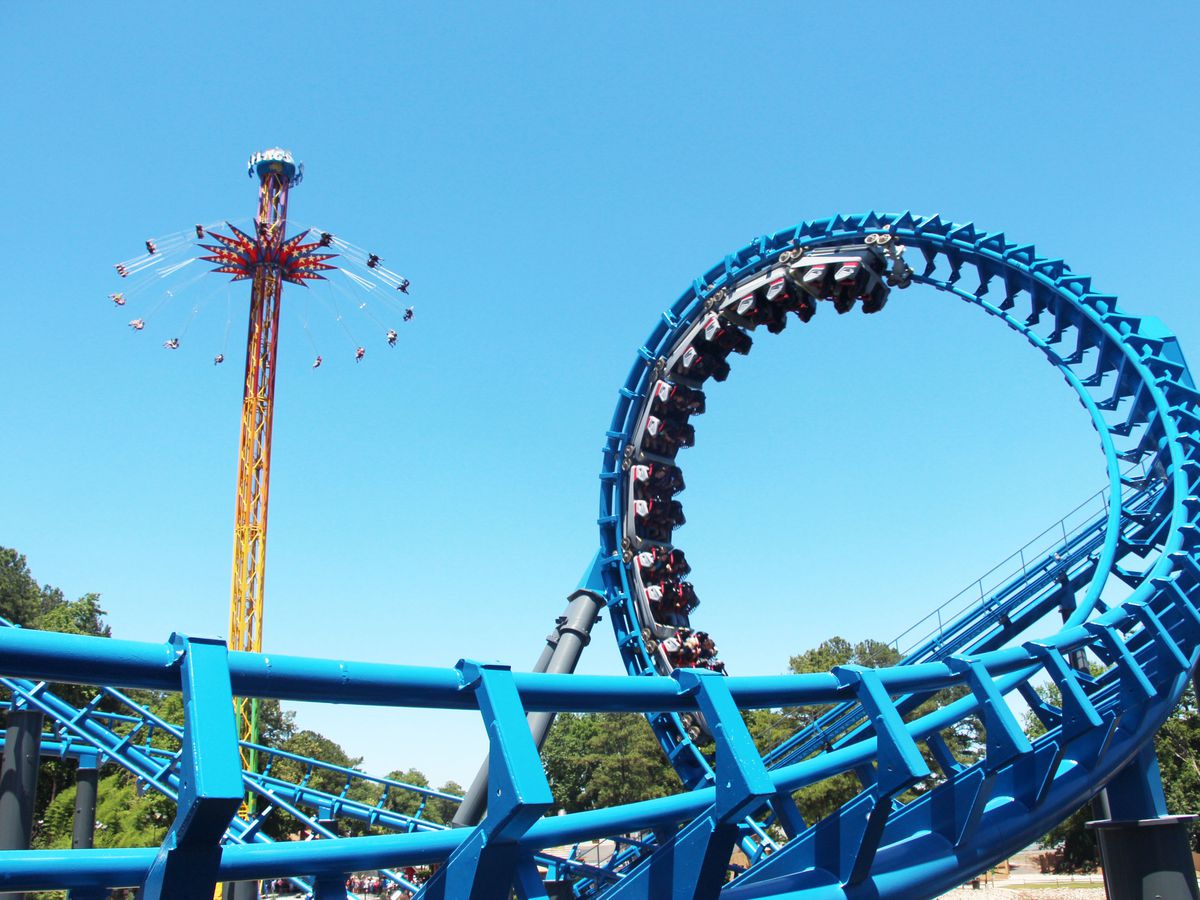A blue rollercoaster with cars full of people traveling on one of the rollercoaster loops. In the distance is another amusement park ride.