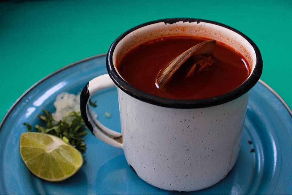 An enamel mug filled with dark red sauce and a clam floating inside, next to lime wedges