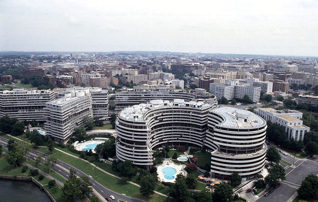 An aerial view of the Watergate hotel. The hotel has an oval shape and a facade with concrete balconies. There is a swimming pool in the central part of the hotel in a courtyard.