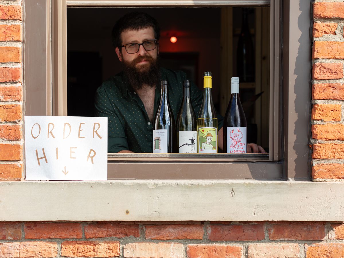 A beard man wearing glasses standing behind four bottles of wine set on a window sill