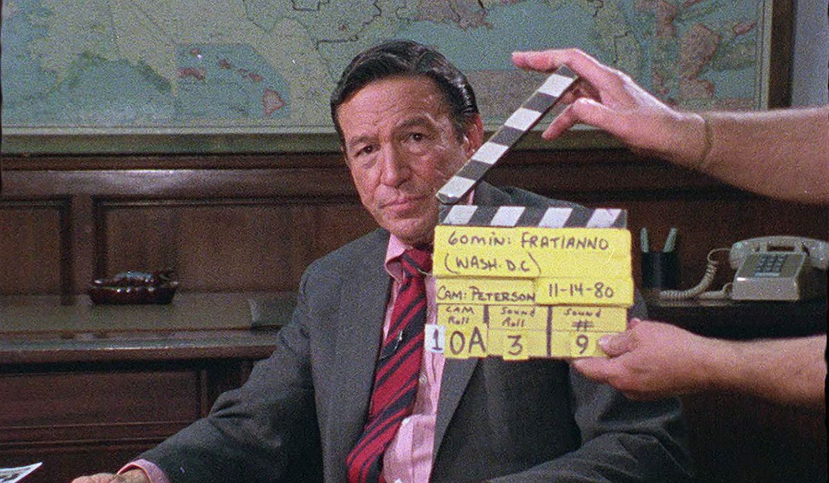 a middle-aged dark-haired man in a pink shirt, red and blue tie, and charcoal jacket — Mike Wallace — with a person holding a clapperboard in front of him in Mike Wallace Is Here